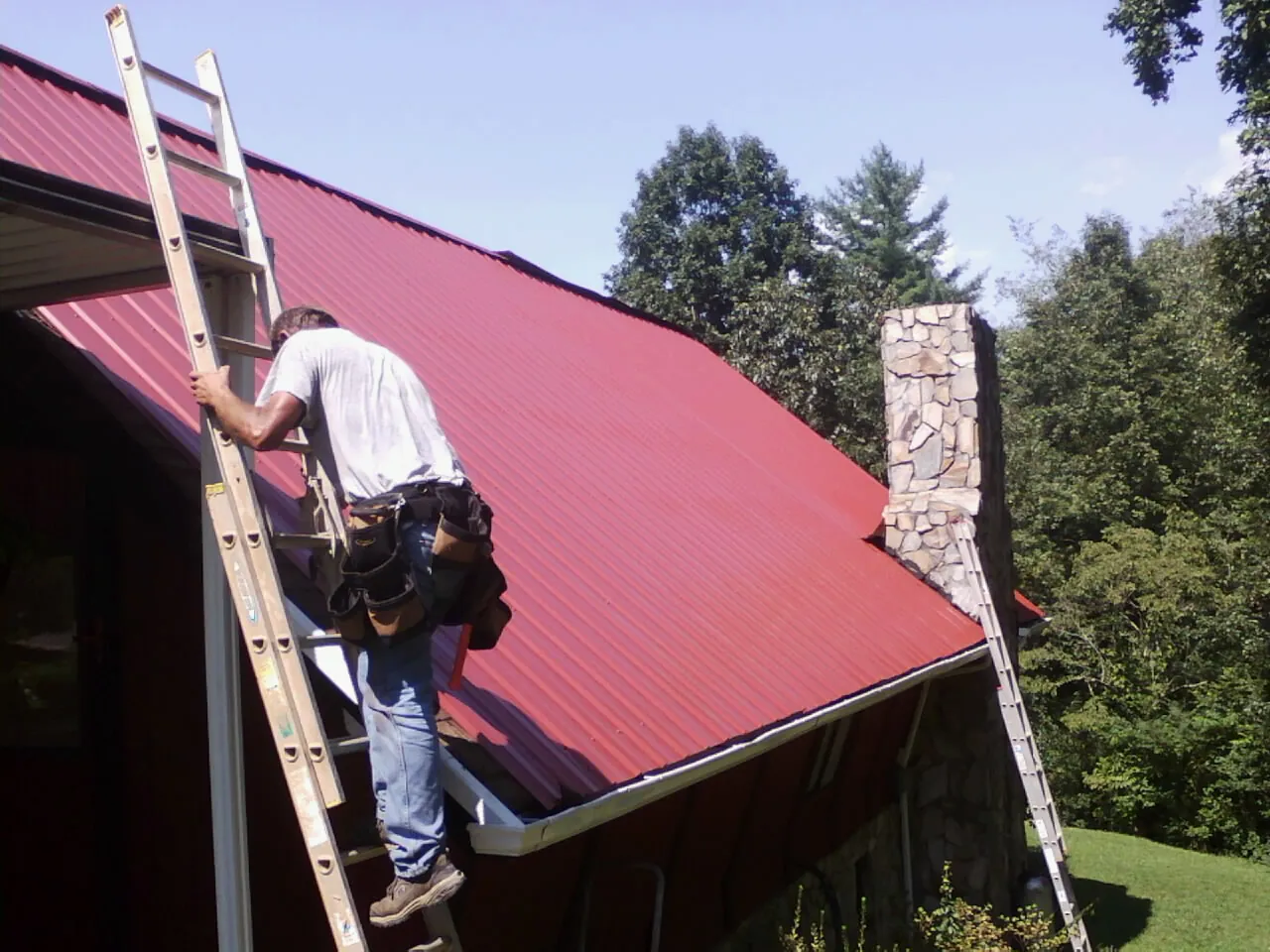 Roofer on a ladder beside a red metal roof and stone chimney — Soffit & Fascia Repair by Roofing Brothers in Caldwell